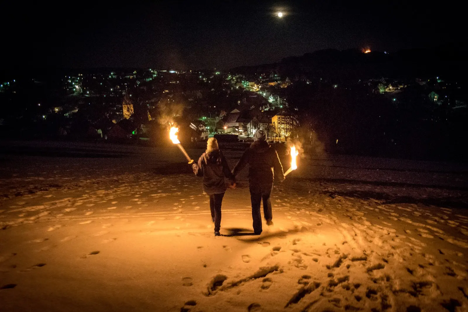 Zwei Menschen gehen mit Fackeln bei Dunkelheit über den Schnee, im Hintergrund die Lichter von Usseln